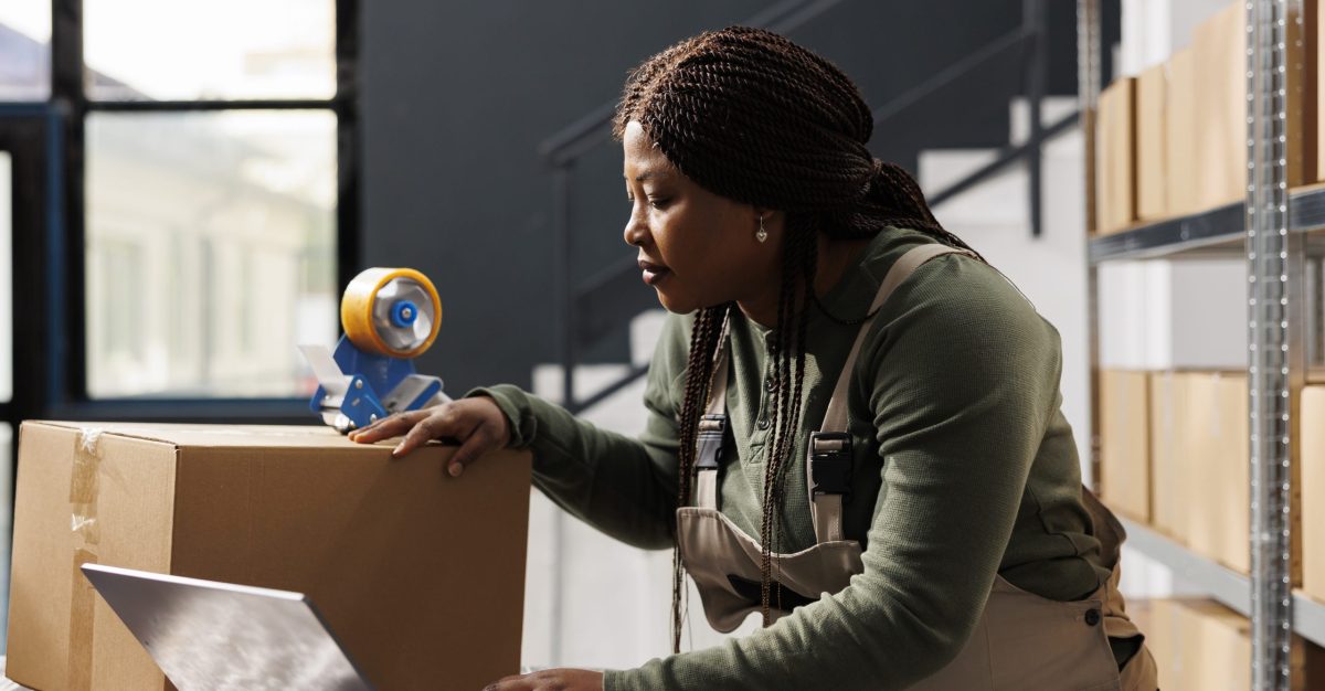 African american worker preparing customers orders using cardboard boxes, checking delivery details on laptop computer in storehouse. Stockroom employee working at products inventory report